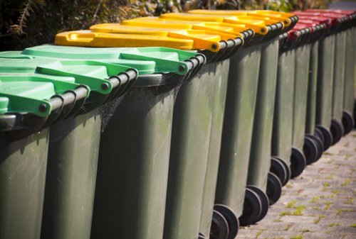 A row of large wheeled rubbish bins situated on a paved pathway, with the front row featuring green-lidded containers, followed by yellow-lidded bins in the middle, and red-lidded bins at the far end. The bins are made of durable plastic with textured surfaces and are positioned closely together, aligned neatly along a curbside. The background includes some greenery and blurred outdoor elements, suggesting an exterior environment likely in Croydon. The image reflects typical waste collection setups used in general rubbish removal services, with the bins arranged for efficient disposal or collection, indicative of a well-maintained residential or community area.