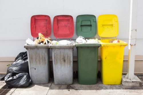 The image displays four wheeled rubbish bins positioned outdoors against a light-colored wall, arranged in a row on a paved surface. From left to right, the first bin is grey with a red lid, containing various discarded items such as paper, plastic wrappers, and other miscellaneous waste spilling slightly over the top. The second bin is also grey with a red lid, similarly filled with refuse including crumpled paper and plastic packaging. The third bin is dark green with a matching green lid, which is closed, and appears to contain waste as no overflow is visible. The fourth bin is bright yellow with a yellow lid; its lid is slightly ajar, revealing some waste inside. To the left of the bins, there are black plastic rubbish bags piled on the ground, partially leaning against the grey bin on the far left. The setting suggests a typical external rubbish collection area, possibly in a residential or commercial context within Croydon or nearby. The arrangement and variety of waste indicate a common bin collection point managed for rubbish disposal, typical of local waste management services in the area served by Garden Clearance Croydon.