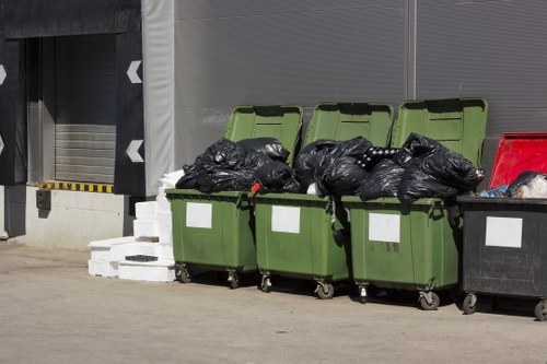A row of four large, green wheeled rubbish bins with open lids, filled with black trash bags, is positioned on a concrete surface in front of a building with grey cladding and a roller shutter door. To the right, there is a larger black bin also filled with rubbish, partially visible. Behind the bins, white concrete blocks are stacked on the ground, and the environment suggests an industrial or commercial area, likely in or near Croydon. The scene illustrates typical waste collection storage, aligning with rubbish removal services by Garden Clearance Croydon, serving local residential and business clients in the area.