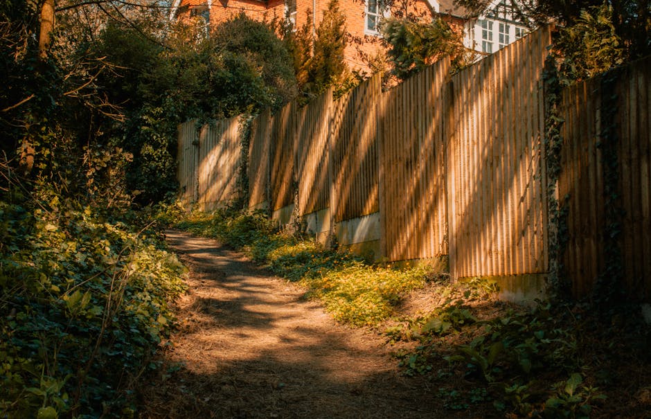 A narrow garden alleyway featuring a dirt and grass pathway flanked by a wooden fence on the left and dense green foliage, including trees and shrubs, on the right. Several wooden pallets are stacked