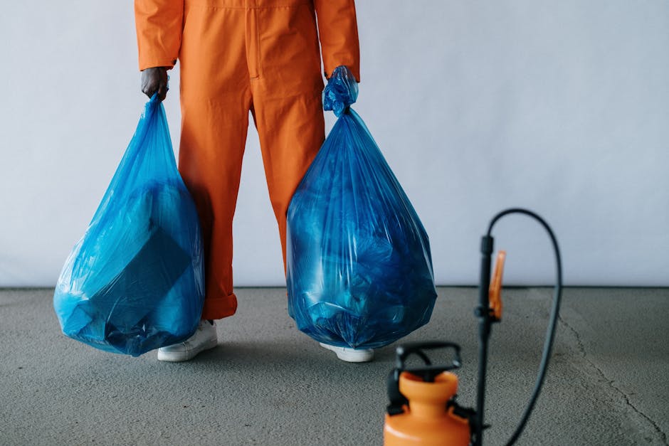 A person dressed in orange protective overalls and white shoes, visible from the waist down, is holding two large blue plastic rubbish bags filled with waste. The bags are semi-transparent, revealing 